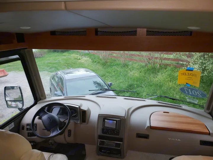Front interior of an RV rental: dashboard, steering wheel, and a clear view of the outside with a car and greenery.. Winnebago Vista 2017