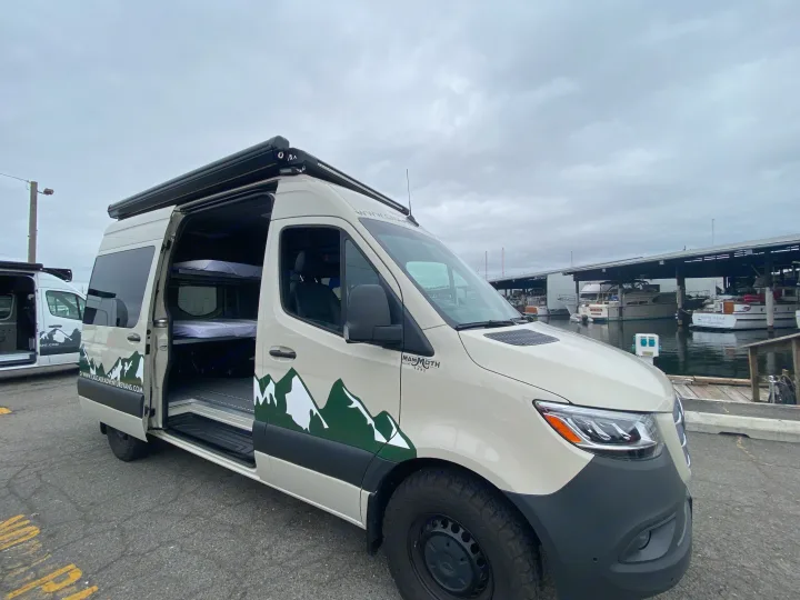 This custom Mammoth adventure van, an RV rental, with bunk beds inside, is parked by boats at a marina.. Mercedes-Benz Sprinter 2023
