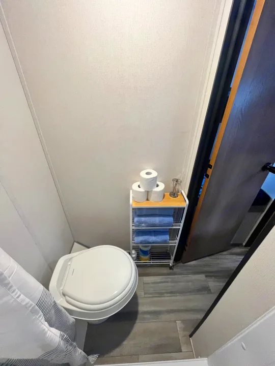 Compact RV rental bathroom with toilet and shelving for essentials like toilet paper and blue towels. Door is open.. Avenger LT 2022