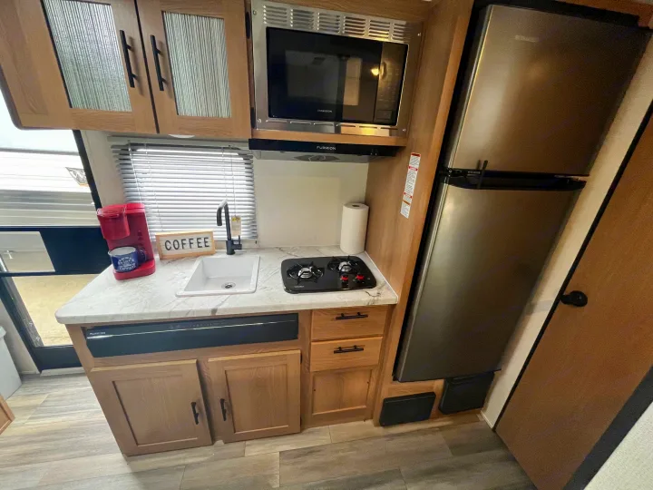 Fully equipped RV rental kitchen: compact layout with wood cabinets, stainless microwave, fridge, sink, and two-burner stove.. Avenger LT 2022