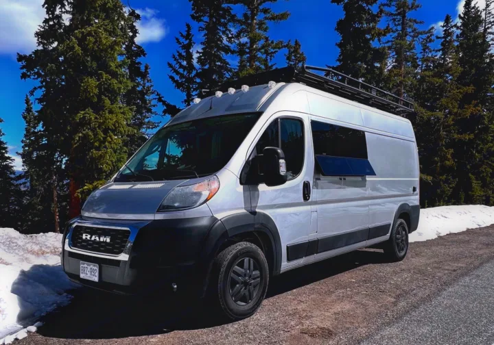 Exploring nature in a white RV rental camper van, parked on a snowy mountain road among tall pine trees.. Ram Promaster 2019