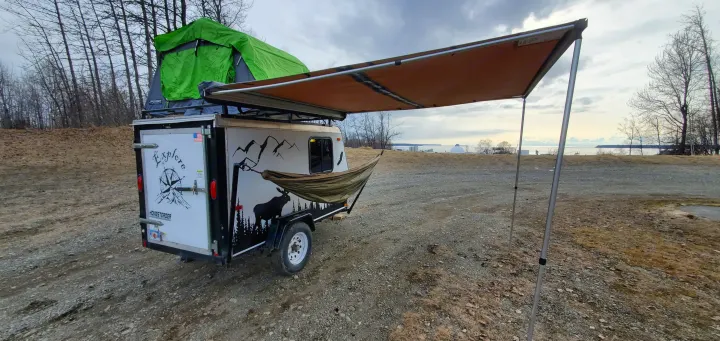 Overland trailer with rooftop tent, hammock, and awning, set up for camping near a body of water—ideal for an RV rental adventure.. Haulmark Homesteader 2005
