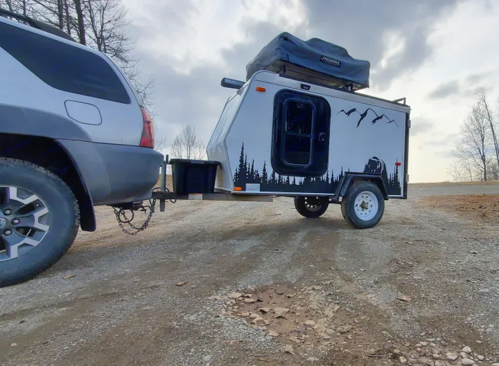 An SUV tows an RV rental teardrop camper with a rooftop tent, ready for outdoor travel on a dirt road.. Haulmark Homesteader 2005