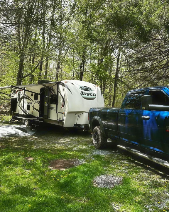 This RV rental features a blue truck hitched to a Jayco camper, parked at a sunny forest campsite.. Jayco Eagle 2016
