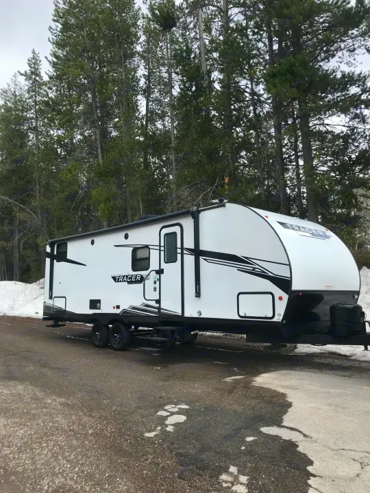 This white Tracer RV rental is parked among tall trees with patches of snow, ready for your next outdoor camping trip.. Forest River Tracer LE 2022