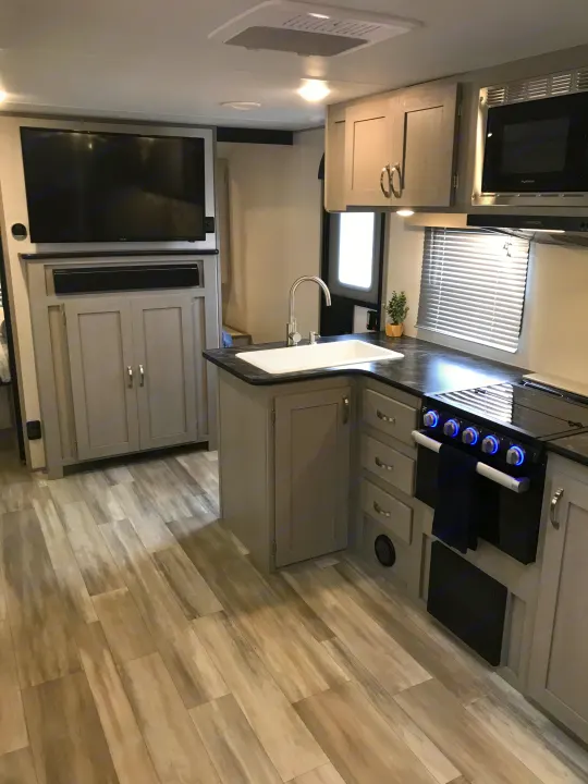 RV rental kitchen and entertainment area: A modern interior with light wood floors, gray cabinets, white sink, black stove, microwave, and a TV.. Forest River Tracer LE 2022