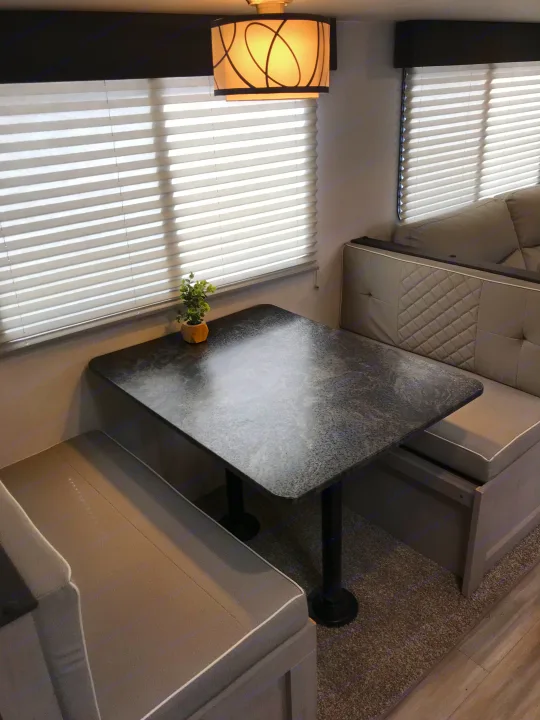 RV rental dinette: a cozy booth with light cushions, dark table, window blinds, and a modern overhead light.. Forest River Tracer LE 2022