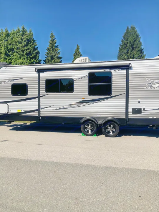 Grey-striped RV rental travel trailer with black windows, parked on asphalt under a blue sky, surrounded by trees.. Jayco Jay Flight SLX Rocky Mountain Edition 2021