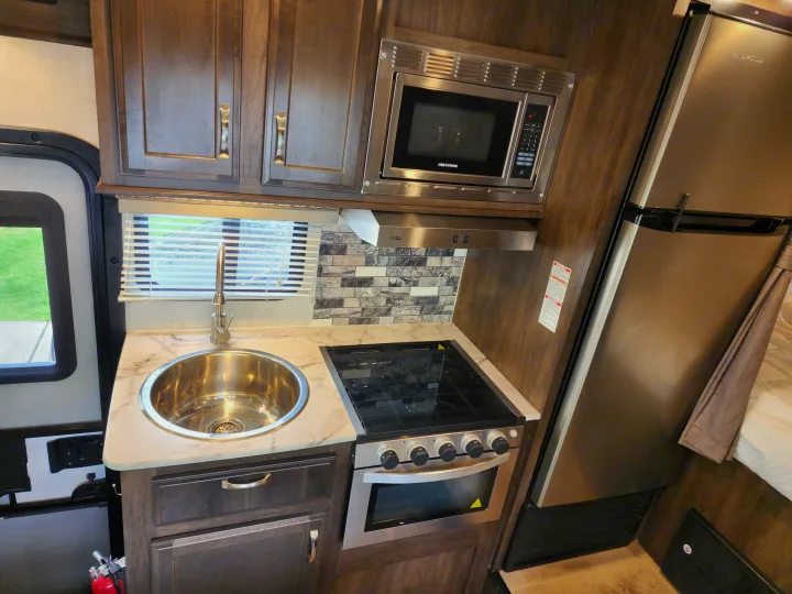 An RV rental kitchenette with a stainless steel microwave, stove/oven, refrigerator, and sink, amidst dark wood cabinets.. Gulf Stream Conquest 2021