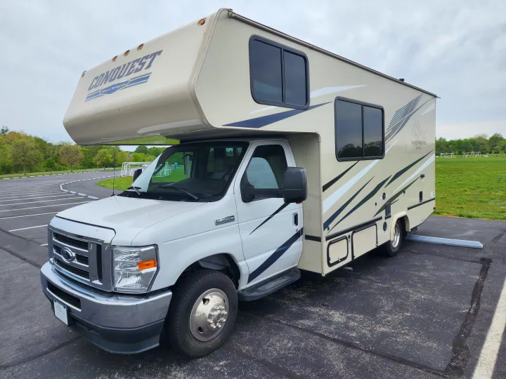 A cream and white Class C Moises Family RV rental, with "Conquest" branding, parked on an asphalt lot near a green field.. Gulf Stream Conquest 2021