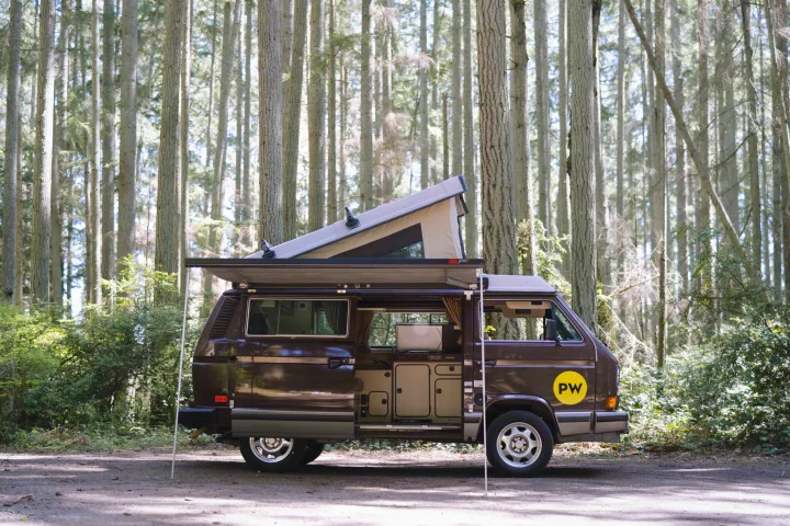 Brown VW camper van with pop-top and awning deployed, ready for adventure in a sunny forest. Perfect RV rental.. Volkswagen Westfalia 1985