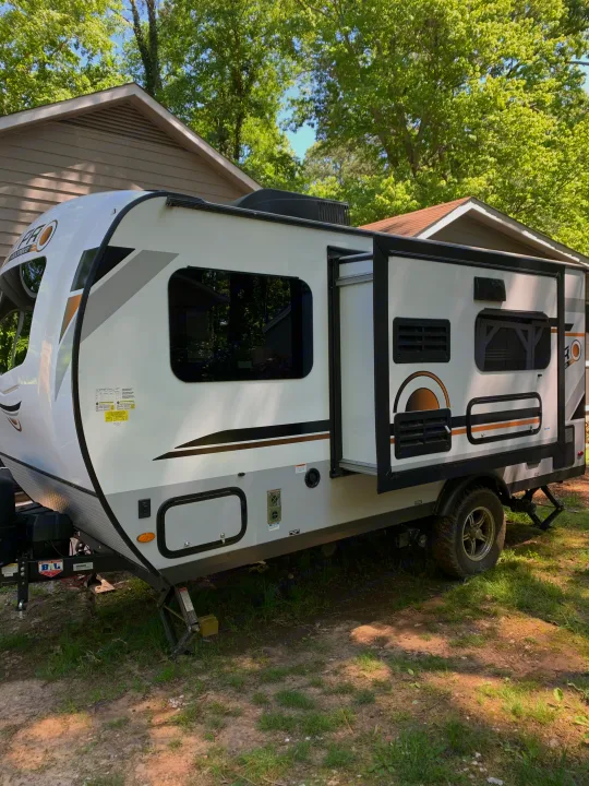 White travel trailer available for rv rental, parked on dirt and grass with buildings & trees in background.. Forest River Rockwood Geo Pro 2020