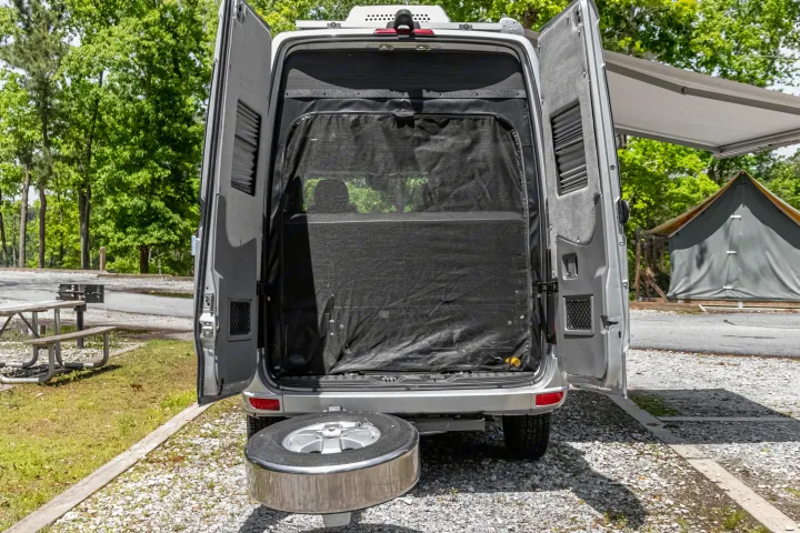 This RV rental shows its open rear with a bug screen and spare tire, at a sunny campsite with trees, picnic table, and tent.. Roadtrek Roadtrek Class B 2017