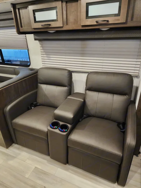 Luxurious RV rental interior featuring recliners with lighted cupholders, wood cabinetry, and window blinds.. Coachmen Leprechaun 2023