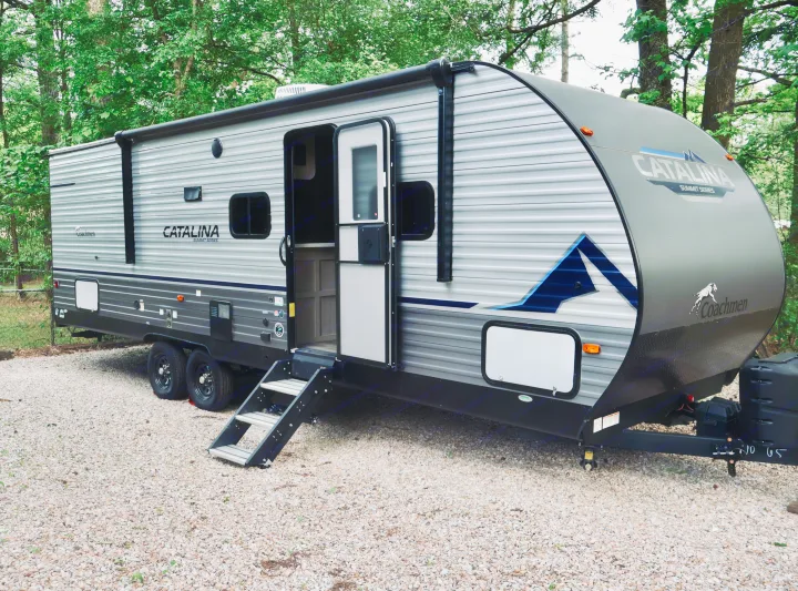 A sleek grey Coachmen Catalina Summit travel trailer, perfect for an RV rental adventure, is parked on gravel among green trees.. Coachmen Other 2023