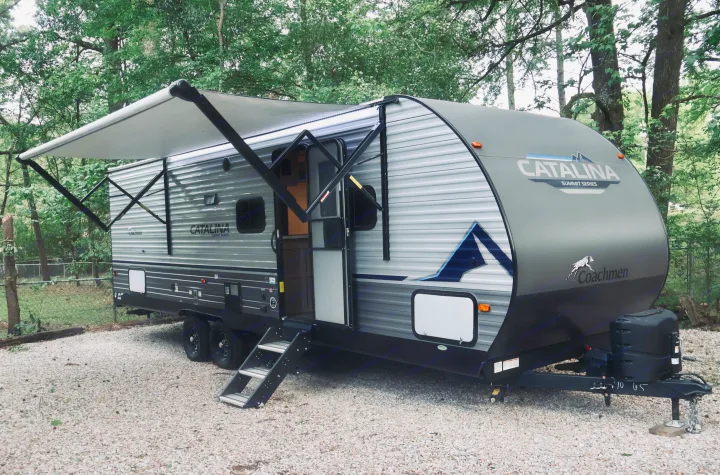 Modern gray travel trailer with extended awning for your next rv rental. Parked on gravel in a lush green wooded campsite.. Coachmen Other 2023