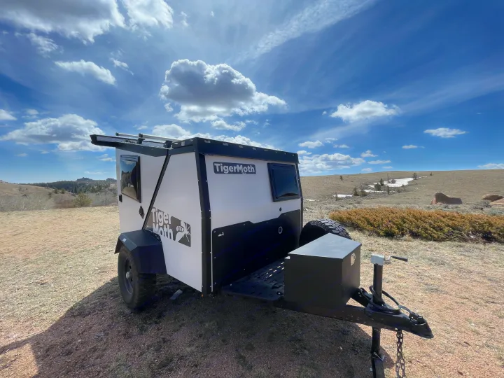White & black TigerMoth compact RV rental parked on a dirt hill with rocky terrain, under a partly cloudy sky.. TAXA Outdoors Tigermoth Camper 2021