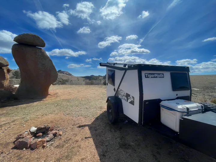 A white and black TigerMoth teardrop RV rental is parked in a dry, open landscape next to a large balanced rock formation and a fire pit under a blue sky.. TAXA Outdoors Tigermoth Camper 2021