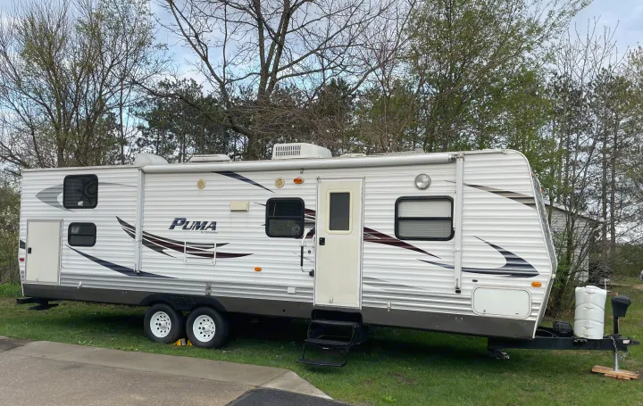 A white "PUMA" travel trailer, ideal for rv rental, parked on grass. Features windows, an awning, and roof units under cloudy skies.. Puma 30DBSS 2009