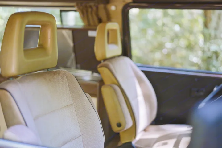 Tan interior of an RV rental, focusing on the textured front seats with distinctive, hollow headrests. Nature visible outside.. Volkswagen Westfalia 1985