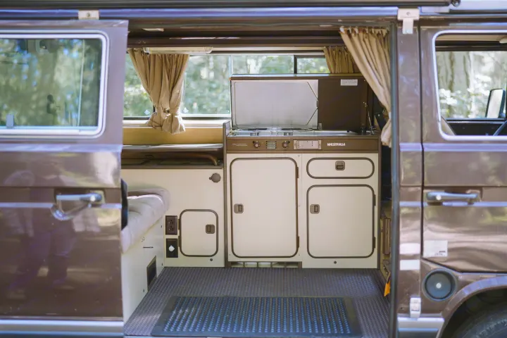 Interior view of a Westfalia RV rental, showing its practical kitchenette with stove, sink, cabinets, and a cozy bench area.. Volkswagen Westfalia 1985