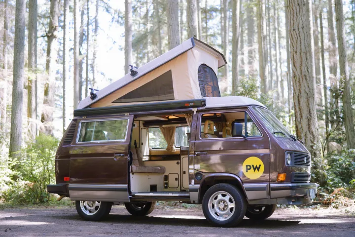 A brown VW pop-top camper van, with its door open revealing the cozy interior, parked in a sunny forest. Perfect for RV rental.. Volkswagen Westfalia 1985