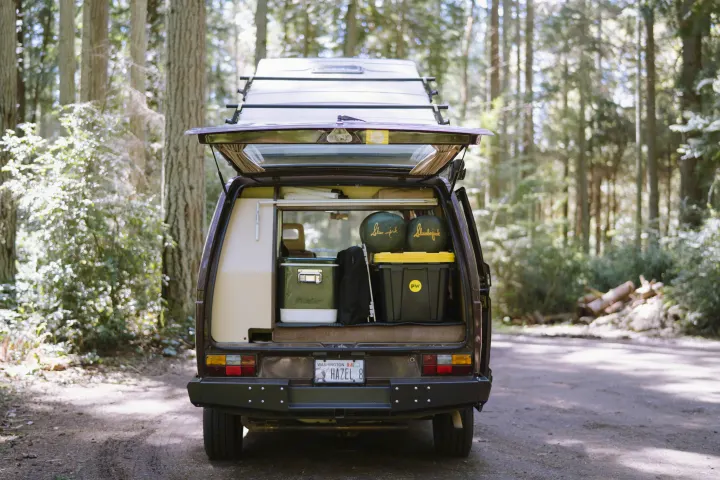 A brown camper van named 'Hazel' (WA plate) with its rear hatch open, loaded with camping gear, parked in a forest. Perfect for an RV rental adventure.. Volkswagen Westfalia 1985