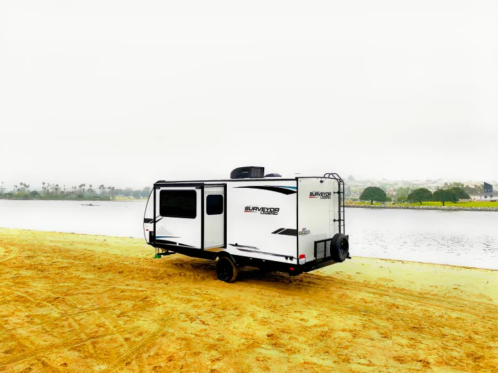 White Surveyor Legend travel trailer for rv rental, parked on a sandy beach next to the water on an overcast day.. Forest River Surveyor Legend 2023
