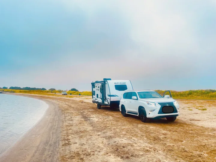 White SUV with an RV rental on a sandy shore by calm water, perfect for outdoor travel.. Forest River Surveyor Legend 2023