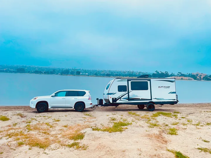 White SUV towing an RV rental (Surveyor Legend) on a sandy shore next to water, with a hazy city skyline.. Forest River Surveyor Legend 2023