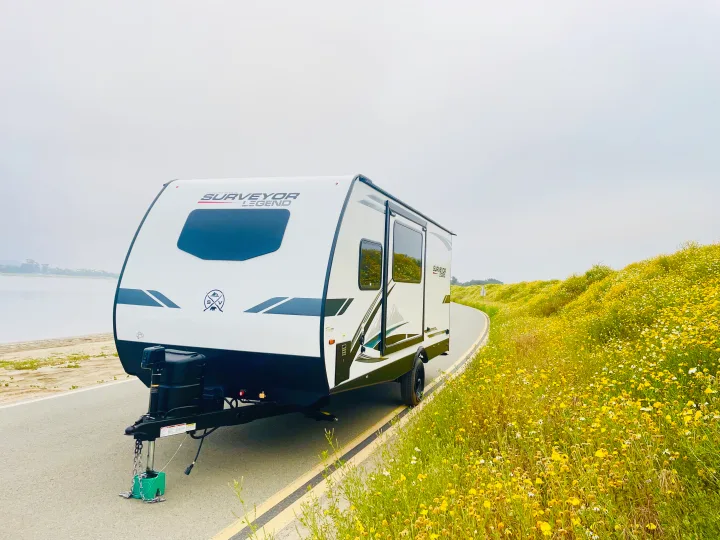 This white Surveyor Legend travel trailer RV rental is parked on a scenic road by a lake and vibrant yellow flower fields.. Forest River Surveyor Legend 2023