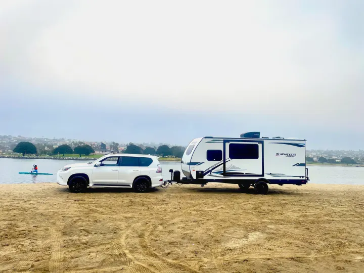 RV rental adventure: White SUV and travel trailer on a sandy beach, with a person paddling on the water.. Forest River Surveyor Legend 2023