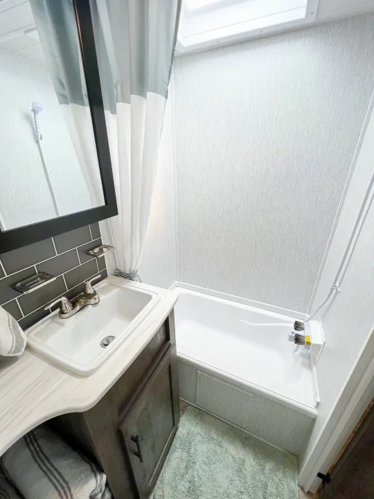 This clean, modern RV rental bathroom features a sink, mirror, and compact tub/shower with a gray curtain.. Forest River Wildwood X-Lite 2022