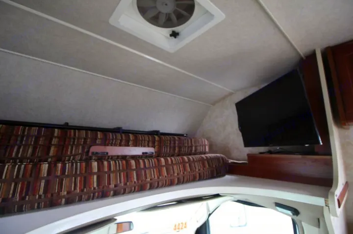 Spacious RV rental cab-over area with comfy striped cushions, wall-mounted TV, and a roof vent for air circulation.. Winnebago Access 2011