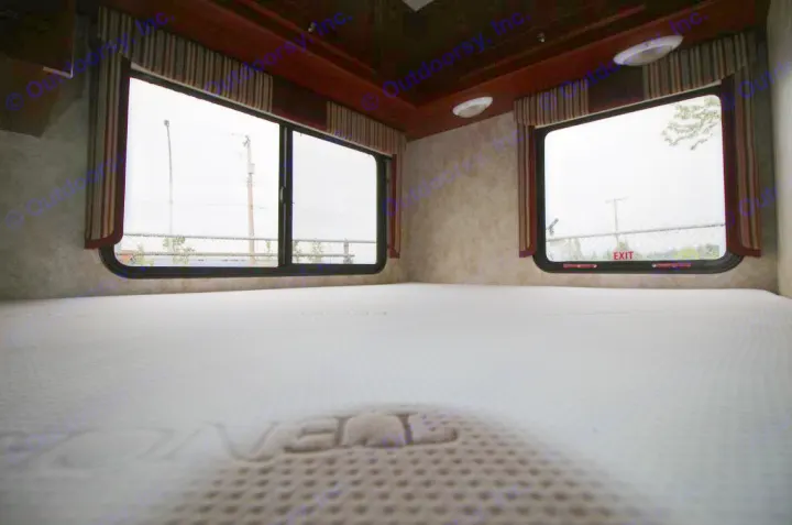 Inside an RV rental bunk: a spacious white mattress, ready for rest. Windows show a rainy outside, with an EXIT sign visible.. Winnebago Access 2011