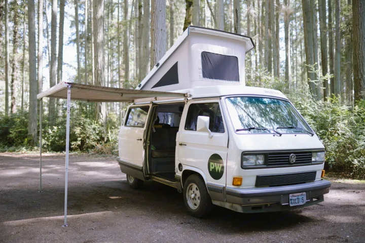 A cozy white VW Westfalia camper van, ideal for rv rental, parked in a lush forest campsite with its pop-top and awning out.. Volkswagen Westfalia 1990