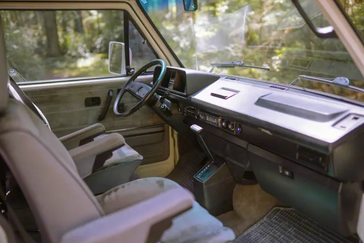Interior of a classic RV rental van: driver's view of the dashboard, steering wheel, and front seats, with a forest outside.. Volkswagen Westfalia 1990