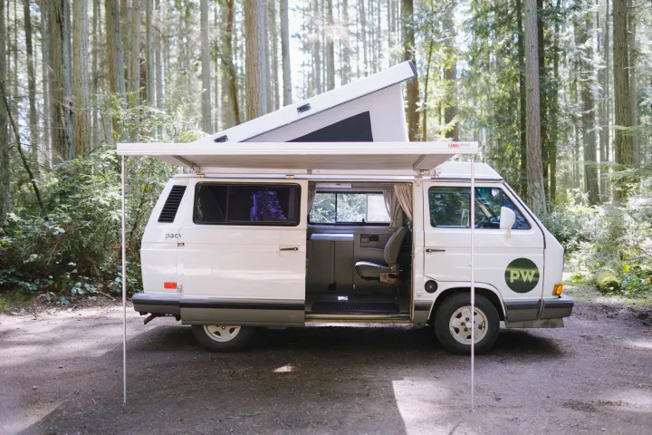 White pop-top camper van (RV rental) with awning extended, parked on a forest path. Side door open, ready for adventure.. Volkswagen Westfalia 1990