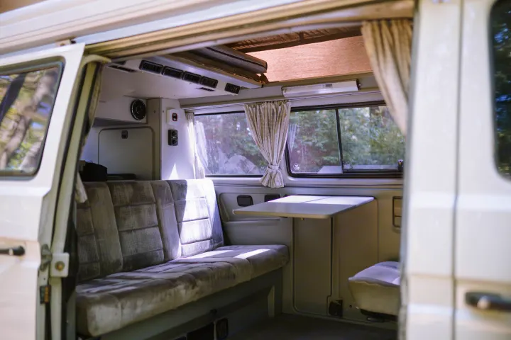 Interior of an RV rental camper van, showing a comfortable bench seat, fold-down table, and windows with curtains, perfect for travel.. Volkswagen Westfalia 1990