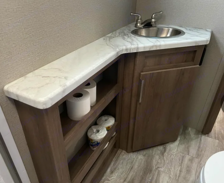 RV rental compact bathroom featuring a corner sink, marble-look countertop, wood vanity with cabinet & open toilet paper storage.. Grand Design Imagine XLS 2021