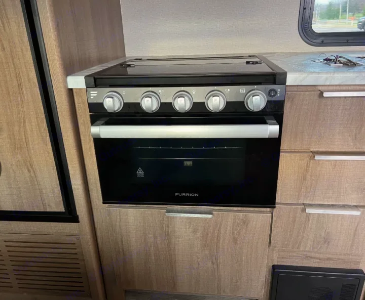 RV rental kitchen: a modern Furrion stove/oven combo with silver controls, set in light wood cabinetry & white countertop.. Grand Design Imagine XLS 2021