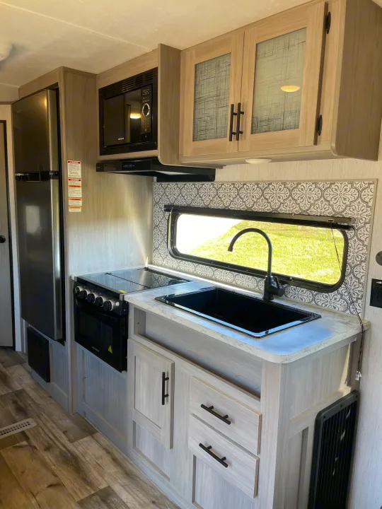 This rv rental features a modern kitchen with light wood cabinets, fridge, microwave, stove, and sink. Patterned backsplash.. Dutchmen Coleman Lantern LT 2022