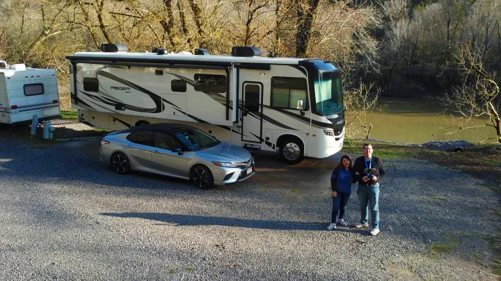 An RV rental and car are parked at a scenic riverside campsite with a travel trailer, a couple stands with a drone remote.. Jayco Precept 2021