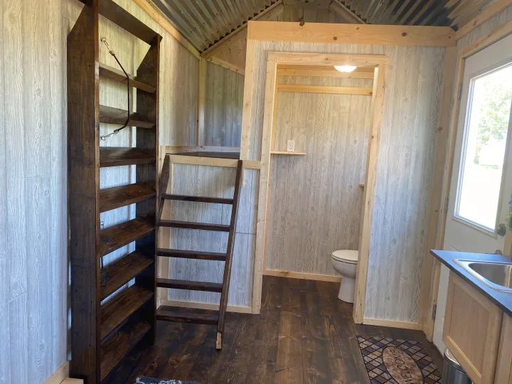 Inside a compact RV rental: dark wood floor, light paneled walls, loft ladder, shelving, bathroom, and kitchen.. Bran Gooseneck 2019