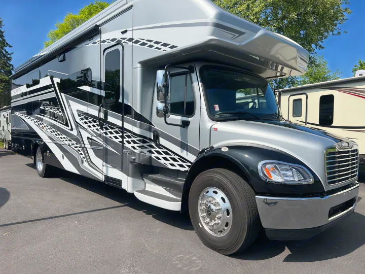 A sleek silver, black, and white motorhome, possibly an RV rental, parked on asphalt with trees in the background.. Entegra Coach Accolade XL 37L  Super-C 2023