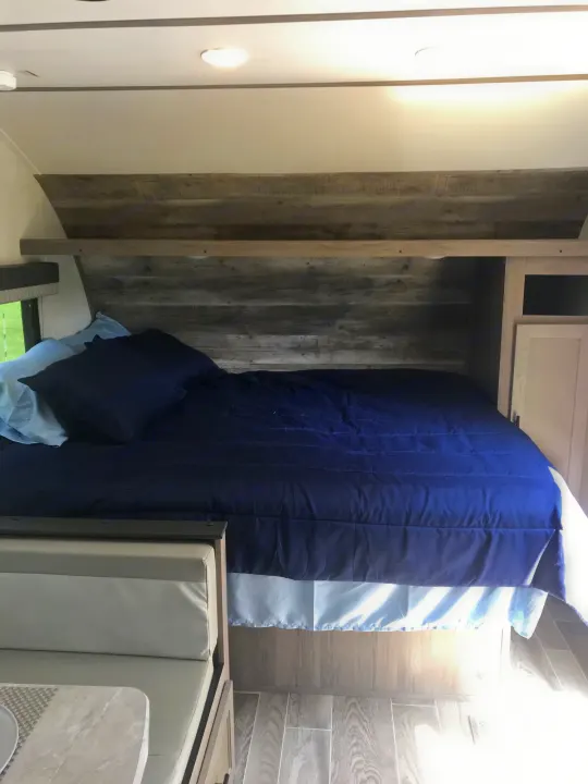 Inside a modern RV rental: A compact bedroom with a blue bed, rustic wood walls, and integrated storage. Dinette visible.. Gulf Stream Friendship 2022