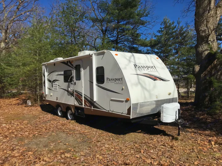 White and brown travel trailer RV parked on a leaf-strewn ground among trees on a sunny day. Ideal RV rental.. Keystone RV Passport Ultra 2013