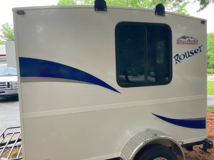 Side view of a white Rouser Runaway RV rental, with blue stripes, a window, and a front-mounted spare tire.. Runaway Rouser 2023