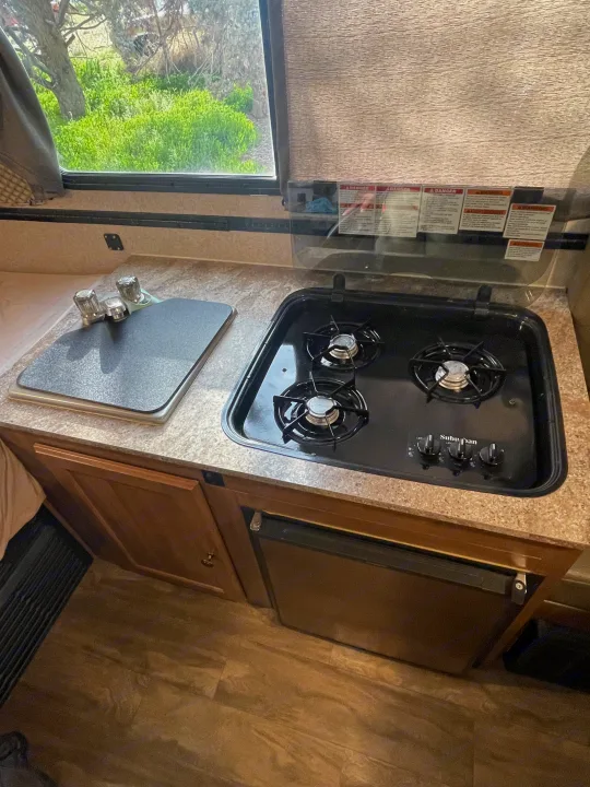 Inside an RV rental kitchen: 3-burner stovetop, covered sink, and under-counter refrigerator. View of green outdoors through window.. Forest River Flagstaff Classic 2019
