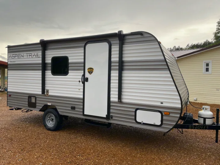 Modern travel trailer for rv rental, featuring striped siding & awning, parked on gravel. Ready for your next adventure!. Dutchmen Aspen Trail 2023